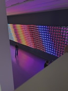 A large, colorful LED light display is mounted on a wall in a spacious room. The display features a variety of vibrant colors in a grid pattern, including blue, red, purple, and yellow. Two people are visible in the room; one is standing and looking at the display, while the other is partially visible, sitting or kneeling closer to the display. The floor is a glossy, reflective surface that matches the colors emanating from the display.