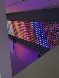 A large, colorful LED light display is mounted on a wall in a spacious room. The display features a variety of vibrant colors in a grid pattern, including blue, red, purple, and yellow. Two people are visible in the room; one is standing and looking at the display, while the other is partially visible, sitting or kneeling closer to the display. The floor is a glossy, reflective surface that matches the colors emanating from the display.