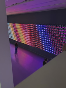 A large, colorful LED light display is mounted on a wall in a spacious room. The display features a variety of vibrant colors in a grid pattern, including blue, red, purple, and yellow. Two people are visible in the room; one is standing and looking at the display, while the other is partially visible, sitting or kneeling closer to the display. The floor is a glossy, reflective surface that matches the colors emanating from the display.