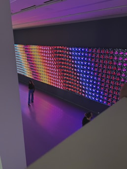 A large, colorful LED light display is mounted on a wall in a spacious room. The display features a variety of vibrant colors in a grid pattern, including blue, red, purple, and yellow. Two people are visible in the room; one is standing and looking at the display, while the other is partially visible, sitting or kneeling closer to the display. The floor is a glossy, reflective surface that matches the colors emanating from the display.
