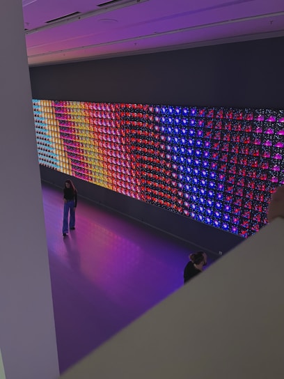 A large, colorful LED light display is mounted on a wall in a spacious room. The display features a variety of vibrant colors in a grid pattern, including blue, red, purple, and yellow. Two people are visible in the room; one is standing and looking at the display, while the other is partially visible, sitting or kneeling closer to the display. The floor is a glossy, reflective surface that matches the colors emanating from the display.