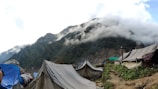 Tents are pitched on a dirt path amidst green foliage on a hillside. The background features mist-laden mountains with clouds rolling over their peaks.