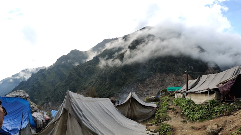 Tents are pitched on a dirt path amidst green foliage on a hillside. The background features mist-laden mountains with clouds rolling over their peaks.