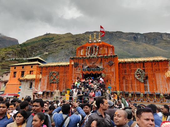 A large temple adorned with vibrant orange marigold decorations and other floral arrangements, set against a backdrop of mountains under a cloudy sky. The scene is bustling with a crowd of people gathered in front of the entrance, some wearing traditional attire, indicating a festive or religious occasion.