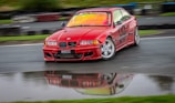 A red BMW car with 'M Performance' and '98' decals is drifting on a wet race track. The car's body is slightly blurred, indicating motion, with a reflection visible in a puddle. The background shows a blurred view of the track and greenery.
