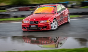 A red BMW car with 'M Performance' and '98' decals is drifting on a wet race track. The car's body is slightly blurred, indicating motion, with a reflection visible in a puddle. The background shows a blurred view of the track and greenery.