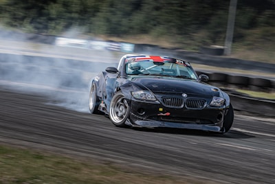 A black sports car is drifting on a race track, generating clouds of smoke from its rear wheels. The car has sponsor decals and a racing number displayed, indicating it is participating in a motorsport event. The background shows a blurred view of the track with tire barriers and greenery, emphasizing the car's speed and motion.