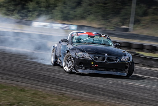 A black sports car is drifting on a race track, generating clouds of smoke from its rear wheels. The car has sponsor decals and a racing number displayed, indicating it is participating in a motorsport event. The background shows a blurred view of the track with tire barriers and greenery, emphasizing the car's speed and motion.