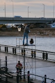 Two workers are involved in an outdoor activity by a river. One worker stands on a scaffold, holding an object and appears to be engaged with a streetlamp. Another worker stands below, near the base of the scaffold. A bridge spans the river in the background with vehicles visible on it, and the scene is set during daylight.