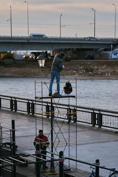 Two workers are involved in an outdoor activity by a river. One worker stands on a scaffold, holding an object and appears to be engaged with a streetlamp. Another worker stands below, near the base of the scaffold. A bridge spans the river in the background with vehicles visible on it, and the scene is set during daylight.