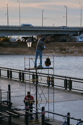 Two workers are involved in an outdoor activity by a river. One worker stands on a scaffold, holding an object and appears to be engaged with a streetlamp. Another worker stands below, near the base of the scaffold. A bridge spans the river in the background with vehicles visible on it, and the scene is set during daylight.