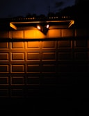 Two bright outdoor lights illuminate a textured metal garage door at night, creating stark contrasts between light and shadow. The warm glow from the lights casts sharp shadows onto the door’s rectangular panels, with a portion of the surrounding darkness partially visible above the door.