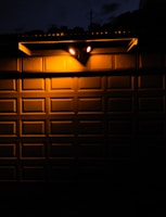 Two bright outdoor lights illuminate a textured metal garage door at night, creating stark contrasts between light and shadow. The warm glow from the lights casts sharp shadows onto the door’s rectangular panels, with a portion of the surrounding darkness partially visible above the door.