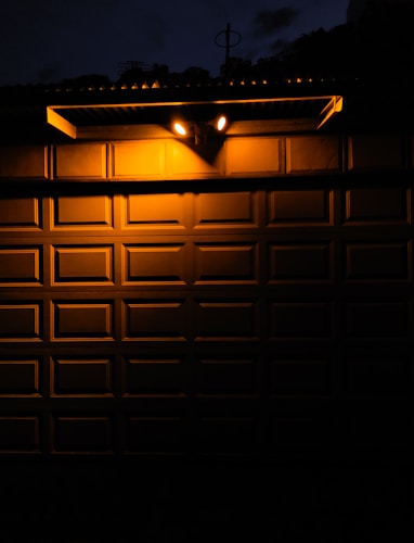 Two bright outdoor lights illuminate a textured metal garage door at night, creating stark contrasts between light and shadow. The warm glow from the lights casts sharp shadows onto the door&rsquo;s rectangular panels, with a portion of the surrounding darkness partially visible above the door.