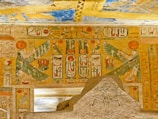A detailed depiction of ancient Egyptian hieroglyphics and art on a stone surface and walls. The imagery includes symbols and figures with wings, likely representing deities or mythical beings, set against a backdrop of vibrant colors and intricate designs.