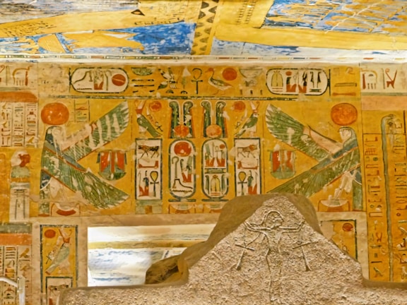 A detailed depiction of ancient Egyptian hieroglyphics and art on a stone surface and walls. The imagery includes symbols and figures with wings, likely representing deities or mythical beings, set against a backdrop of vibrant colors and intricate designs.