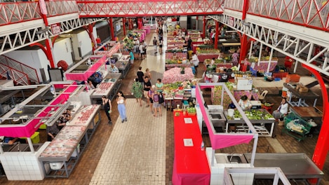 An indoor market with multiple stalls selling various goods including fruits, vegetables, and meats. The market features red and white structures with vendors and customers interacting. The floor is tiled in tones of brown and beige, creating a clean and organized atmosphere.