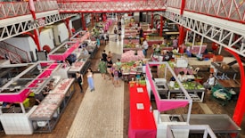 An indoor market with multiple stalls selling various goods including fruits, vegetables, and meats. The market features red and white structures with vendors and customers interacting. The floor is tiled in tones of brown and beige, creating a clean and organized atmosphere.
