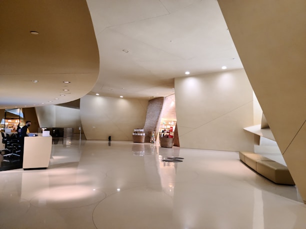 A spacious, modern interior with smooth, curved walls and ceilings. The floor is shiny and reflective, creating a sense of openness. Soft lighting illuminates the space, and a small reception desk is situated on the left, staffed by an individual. In the background, there are shelves with products and a small corridor leading to another area.