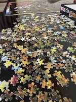 Scattered jigsaw puzzle pieces spread across a dark surface, with an open puzzle box visible in the corner. The pieces display various colors and patterns, suggesting a vibrant image once assembled. Some edges of the puzzle are partially completed, indicating the beginning of assembly.
