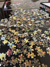 Scattered jigsaw puzzle pieces spread across a dark surface, with an open puzzle box visible in the corner. The pieces display various colors and patterns, suggesting a vibrant image once assembled. Some edges of the puzzle are partially completed, indicating the beginning of assembly.