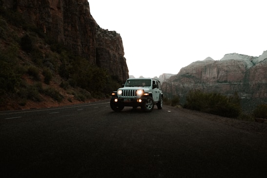 A white Jeep is parked on a winding mountain road surrounded by rocky cliffs and sparse vegetation. The headlights of the Jeep are on, and the surrounding environment appears rugged and serene with tall rock formations in the background.
