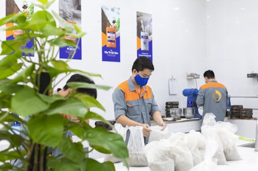 Three workers wearing uniforms with orange and gray colors are performing tasks in a laboratory or industrial setting. One worker is in the foreground handling plastic bags filled with a gray substance, possibly a powder. There's a plant in the foreground on the left, and posters with technical content are on the wall. Equipment and tools are visible on the tables.