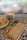 A cup of latte art coffee, a sesame-topped pastry on a white plate, and a book titled 'Las Siete Leyes Espirituales del Éxito' by Deepak Chopra are placed on a patterned, tiled table. The background shows a foggy, possibly rainy window.
