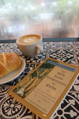 A cup of latte art coffee, a sesame-topped pastry on a white plate, and a book titled 'Las Siete Leyes Espirituales del Éxito' by Deepak Chopra are placed on a patterned, tiled table. The background shows a foggy, possibly rainy window.