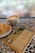 A cup of latte art coffee, a sesame-topped pastry on a white plate, and a book titled 'Las Siete Leyes Espirituales del Éxito' by Deepak Chopra are placed on a patterned, tiled table. The background shows a foggy, possibly rainy window.