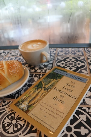 A cup of latte art coffee, a sesame-topped pastry on a white plate, and a book titled 'Las Siete Leyes Espirituales del &Eacute;xito' by Deepak Chopra are placed on a patterned, tiled table. The background shows a foggy, possibly rainy window.