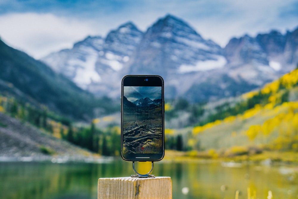 a black smartphone on a wooden post in front of a lake and mountains
