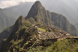 Machu Picchu, an ancient Incan citadel, is perched high on a mountain ridge surrounded by lush, forested peaks and a cloudy sky. Stone structures and terraces are clearly visible, set against the dramatic backdrop of the Andean mountains.