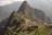 Machu Picchu, an ancient Incan citadel, is perched high on a mountain ridge surrounded by lush, forested peaks and a cloudy sky. Stone structures and terraces are clearly visible, set against the dramatic backdrop of the Andean mountains.