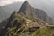 Machu Picchu, an ancient Incan citadel, is perched high on a mountain ridge surrounded by lush, forested peaks and a cloudy sky. Stone structures and terraces are clearly visible, set against the dramatic backdrop of the Andean mountains.