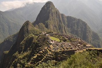 Machu Picchu, an ancient Incan citadel, is perched high on a mountain ridge surrounded by lush, forested peaks and a cloudy sky. Stone structures and terraces are clearly visible, set against the dramatic backdrop of the Andean mountains.