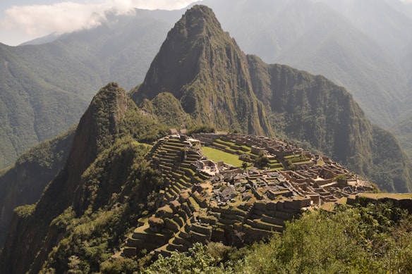 Machu Picchu, an ancient Incan citadel, is perched high on a mountain ridge surrounded by lush, forested peaks and a cloudy sky. Stone structures and terraces are clearly visible, set against the dramatic backdrop of the Andean mountains.