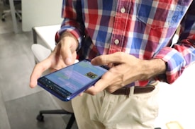 A person wearing a red, blue, and white plaid shirt, holding a smartphone. The phone screen displays a messaging application. The background shows an office environment with desks and chairs.