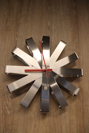 A modern wall clock features an abstract starburst design with metallic rectangular spokes radiating from a central axis. The clock hands are bright red and contrast sharply against the brushed silver background. The clock is mounted on a wooden surface, adding a touch of warmth to the contemporary design.