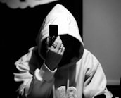 A person wearing a hooded sweatshirt holds a flip phone up to their face, obscuring their identity. The setting appears dimly lit, enhancing the shadowy atmosphere. The person's hand is adorned with rings, adding a touch of style or mystery.