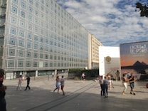 A group of people dance in an open square near a modern building with a grid-like facade. There is a large advertisement featuring pyramids to the right. The scene is set under a partly cloudy sky.