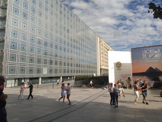 A group of people dance in an open square near a modern building with a grid-like facade. There is a large advertisement featuring pyramids to the right. The scene is set under a partly cloudy sky.