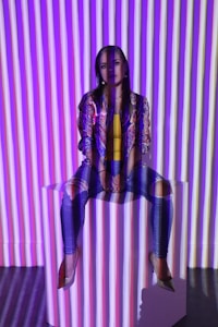 A person is sitting on a white platform with multicolored vertical stripes projected onto them and the background. They appear calm, wearing a patterned jacket, yellow top, and ripped jeans.