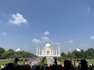 The image depicts a large crowd of people visiting a famous white marble mausoleum with a large dome and minarets on a clear day with a blue sky and a few clouds. The structure is surrounded by lush green gardens and trees.