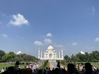 The image depicts a large crowd of people visiting a famous white marble mausoleum with a large dome and minarets on a clear day with a blue sky and a few clouds. The structure is surrounded by lush green gardens and trees.