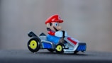 A toy character with a red hat and blue overalls driving a small go-kart. The go-kart has a metallic body and yellow-rimmed wheels.