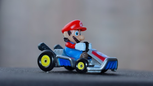 A toy character with a red hat and blue overalls driving a small go-kart. The go-kart has a metallic body and yellow-rimmed wheels.