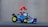 A toy character with a red hat and blue overalls driving a small go-kart. The go-kart has a metallic body and yellow-rimmed wheels.