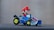 A toy character with a red hat and blue overalls driving a small go-kart. The go-kart has a metallic body and yellow-rimmed wheels.
