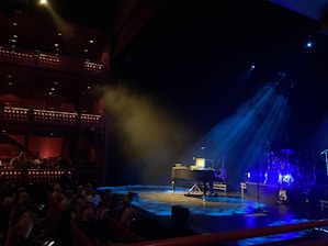 A dimly lit stage is set up for a music performance. A grand piano is placed centrally, accompanied by microphones and guitars on stands. The lighting casts a blue hue over the instruments and creates a dramatic atmosphere. In the foreground, an audience is seated, awaiting the show. The theater's interior features ornate balcony railings and is warmly lit, contributing to a cozy ambiance.